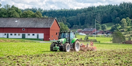 En grön traktor plöjer en åker framför en röd ladugård på landsbygden med skog i bakgrunden.