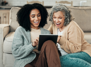 Två kvinnor sitter på golvet framför en soffa och tittar glatt på en surfplatta tillsammans.