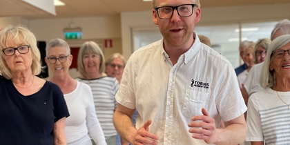 En grupp äldre personer dansar tillsammans inomhus, med en man i vit skjorta i förgrunden.