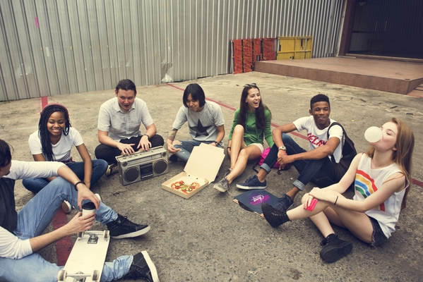 Några unga vuxna sitter på marken utan för en industrilokal och pratar, äter pizza och lyssnar till musik.