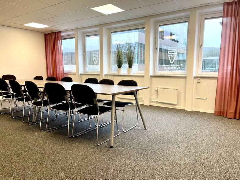 Ett modernt konferensrum med långbord, svarta stolar, stora fönster och röda gardiner.