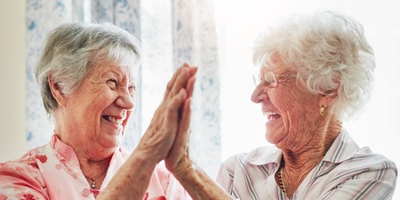 Två äldre kvinnor skrattar och ger varandra en high five i ett ljust rum.