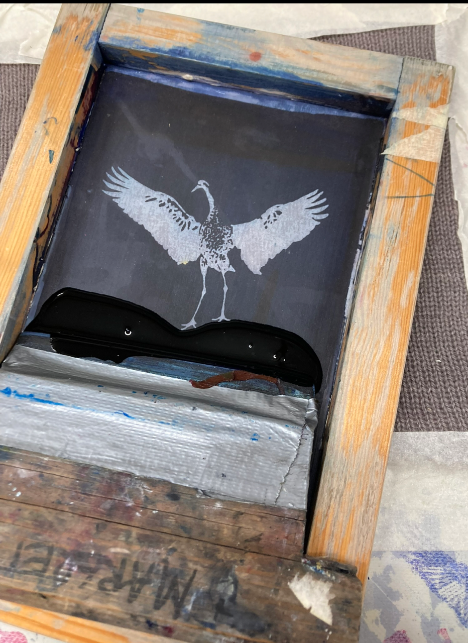 En silkscreenram med en schablon av en trana och svart tryckfärg redo för screentryck.