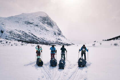 Fyra personer med pulkor står på skidor i ett snötäckt fjällandskap med berg i bakgrunden.
