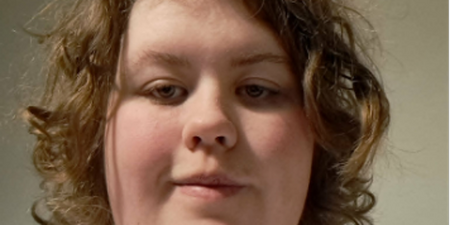 Person med lockigt, blont hår och vit t-shirt sitter framför en neutral bakgrund. Ansiktet är suddat.