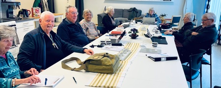 En grupp äldre personer sitter runt ett konferensbord i ett ljust mötesrum med datorer och anteckningar.