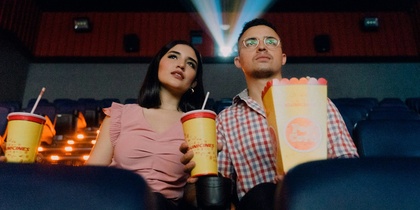 Två personer sitter i en biosalong med popcorn och läsk, filmprojektor lyser bakom dem.