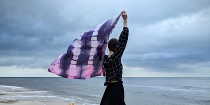 Person står på en omkullfallen trädstam vid havet och håller upp en lila sjal mot himlen.