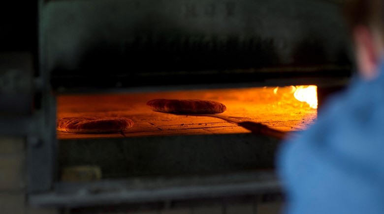 Bröd gräddas i en vedeldad ugn med öppen låga, en person syns suddigt i förgrunden.