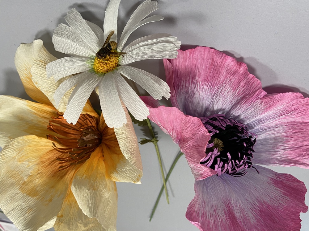 Tre färgglada pappersblommor och en konstgjord bi på ett vitt underlag.