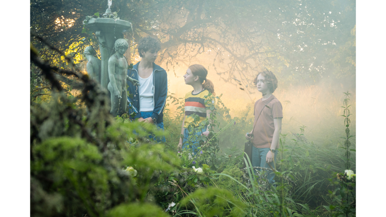 Tre barn står i en dimmig, grönskande trädgård och tittar på en staty.