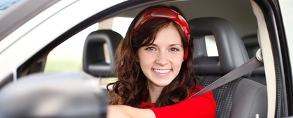 Ung kvinna med långt brunt hår och röd tröja sitter leende bakom ratten i en bil.