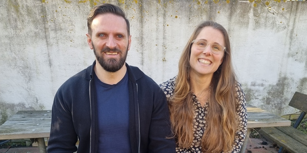 Två personer sitter på en bänk utomhus med en svart ledarhund mellan sig.