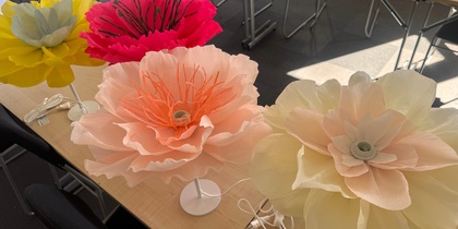 Fyra stora, färgglada konstgjorda blommor på lampfötter står på ett bord i ett mötesrum.