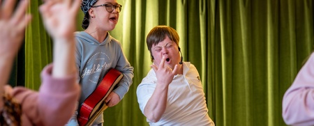 Två unga personer, en med gitarr, sjunger och gör rockposer framför gröna gardiner.