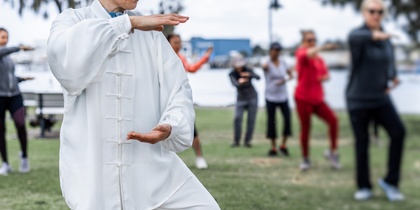 En grupp människor utövar tai chi utomhus i en park, med en kvinna i vit dräkt i förgrunden.