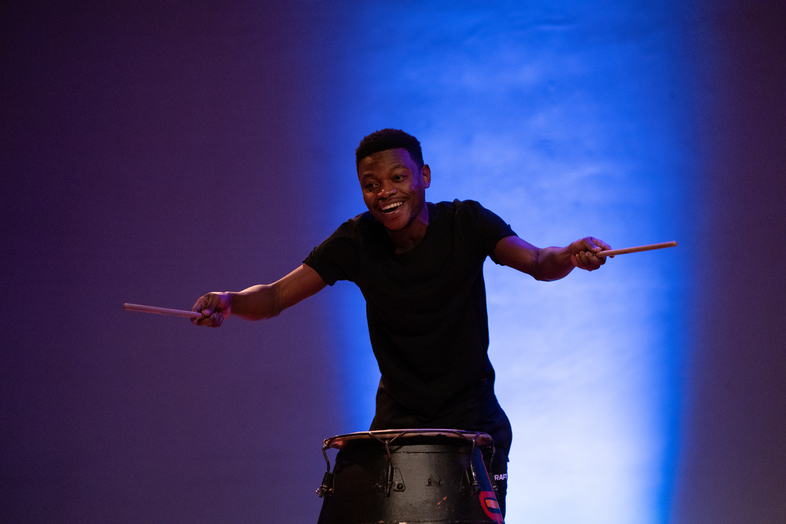 En ung man spelar trumma med trumpinnar på en scen med blå bakgrundsbelysning.