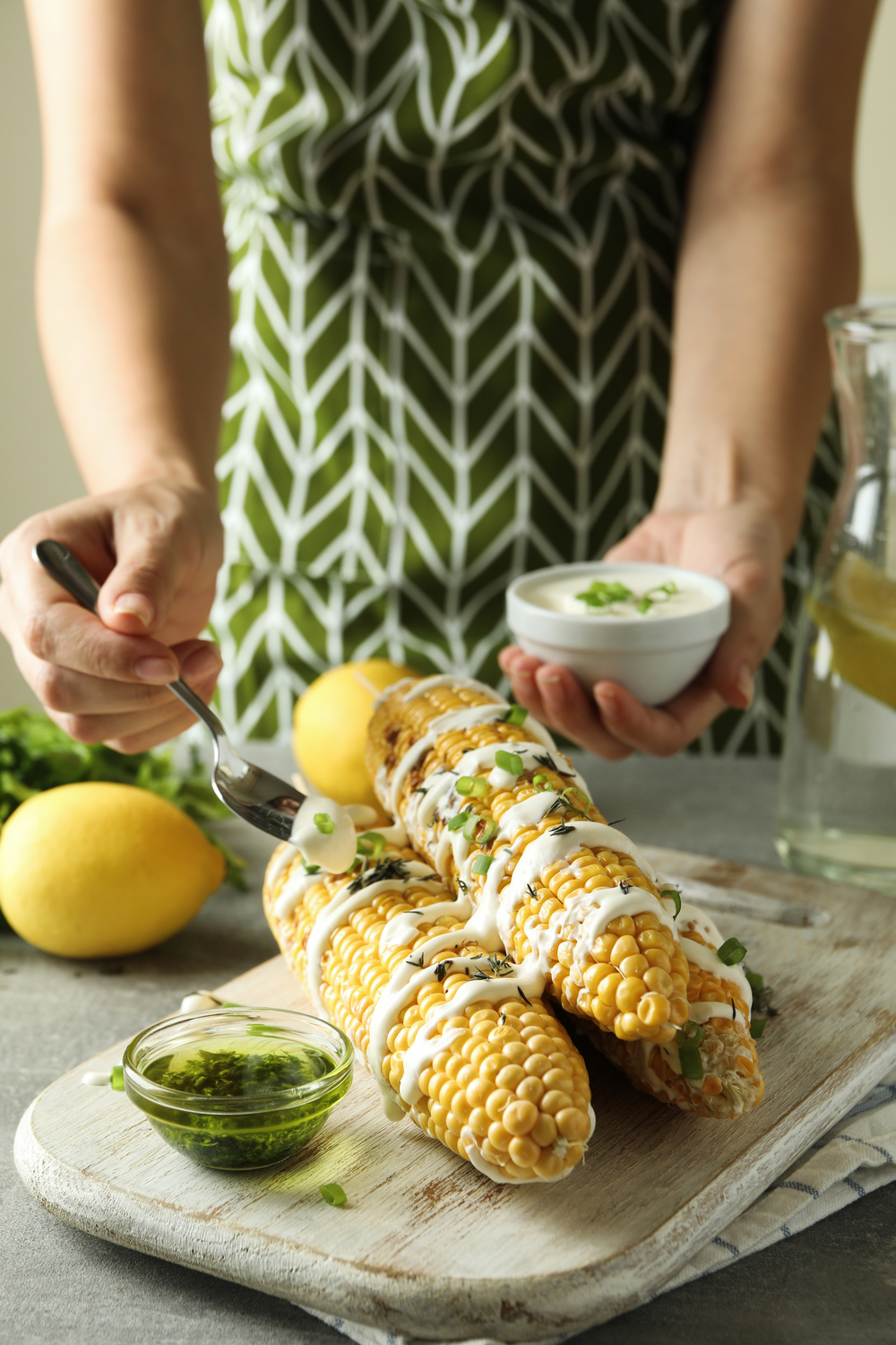 Person dekorerar majskolvar med sås och örter på en skärbräda, citroner och en skål med sås bredvid.