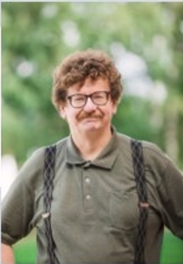 Man med glasögon, mustasch och lockigt hår bär grönt pikétröja och hängslen, står utomhus med suddig grön bakgrund.