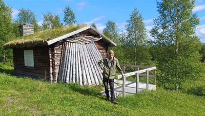 En person står framför en liten timmerstuga med grästak i en grönskande skogsmiljö.