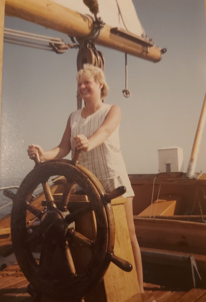 En kvinna styr ett stort trähjul på en segelbåt under en solig dag.