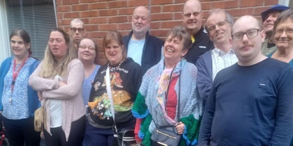 En grupp människor står och sitter utomhus framför en tegelvägg, några med färgglada kläder.