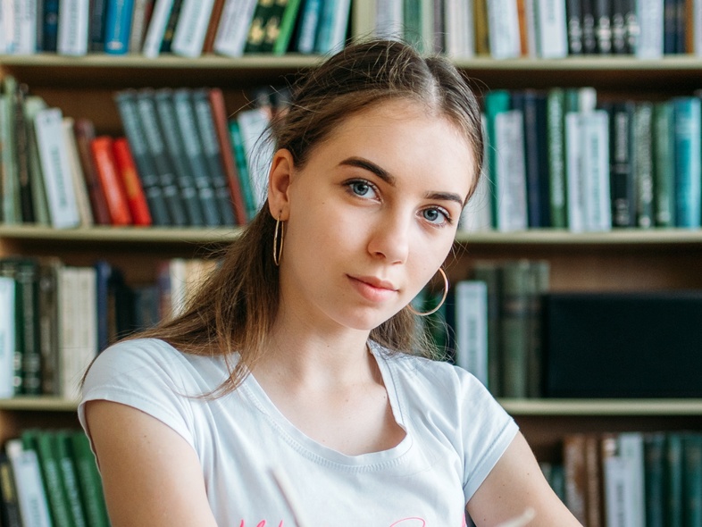 Ung kvinna sitter vid ett bord med böcker i ett bibliotek och tittar mot kameran.