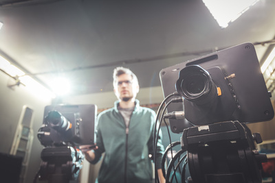 En person står suddigt i bakgrunden bakom två filmkameror i en studio med stark belysning.