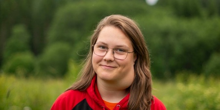 Ung person med glasögon och röd-svart hoodie står utomhus framför en grönskande skog.