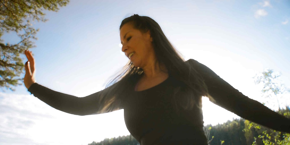 Person i svart tröja står utomhus med utsträckta armar mot en ljus himmel och skog i bakgrunden.