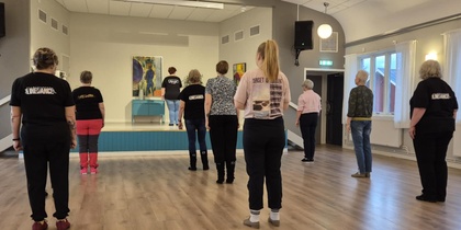 En grupp personer står i ett dansrum och följer en instruktör på en liten scen längst fram.
