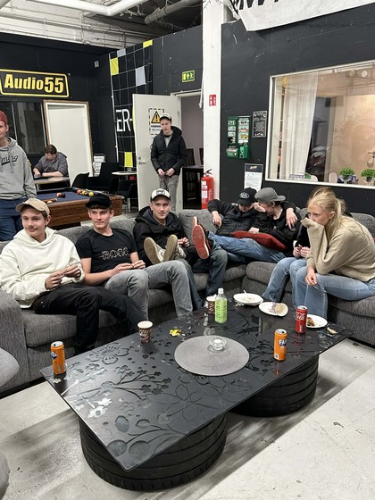En grupp unga vuxna sitter i en soffa runt ett svart soffbord med drycker och tallrikar i ett vardagsrumsliknande rum.