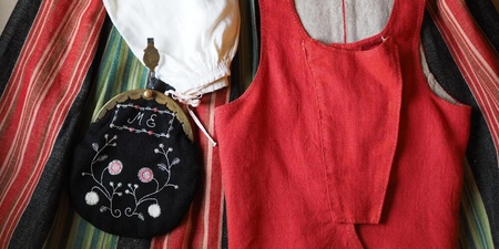 Traditionella klädesplagg inklusive en röd väst, en vit mössa och en broderad svart väska på randigt tyg.