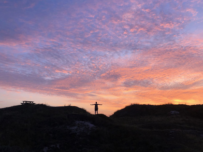 Person står med utsträckta armar på en kulle vid solnedgång, himlen är färgad i rosa och lila nyanser.