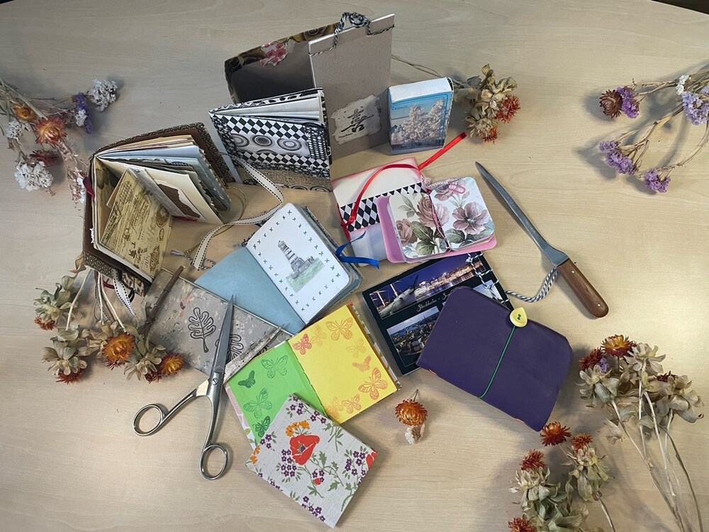 Ett bord med handgjorda anteckningsböcker, sax, brevkniv och torkade blommor utspridda.