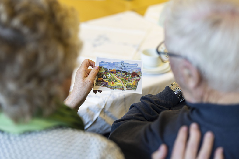 Två äldre personer sitter tillsammans och tittar på ett färgglatt vykort vid ett dukat bord.