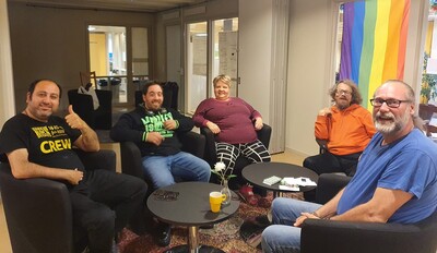 Fem personer sitter i en lounge och samtalar, med en regnbågsflagga i bakgrunden.