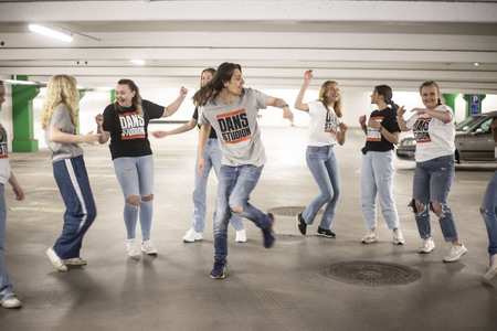En grupp unga personer dansar tillsammans i ett parkeringsgarage, alla bär jeans och t-shirts med texten 'DANS STUDIO'.