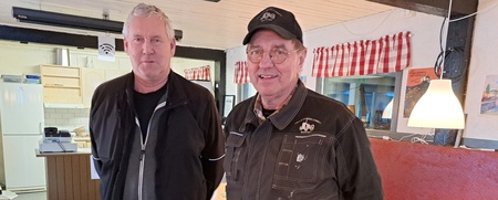 Två män står i ett enkelt inrett rum med träbord och stolar, båda klädda i mörka arbetskläder.