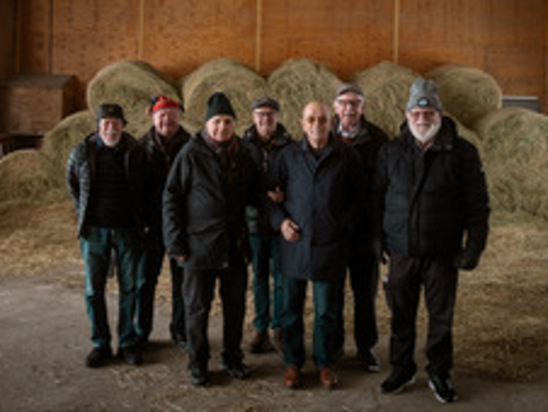 Sju äldre män står tillsammans i en lada framför staplade höbalar.