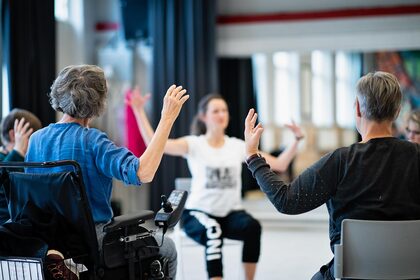 En grupp äldre personer deltar i sittande rörelseövningar i en ljus sal, ledda av en instruktör.