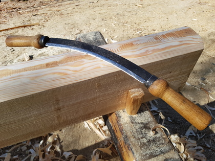 En dragkniv med två trähandtag ligger på en trästock utomhus, omgiven av träspån.