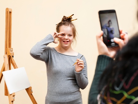 Ung kvinna med penslar i håret poserar framför ett staffli medan någon tar ett foto med mobiltelefon.