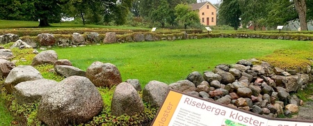 Stenmur och grön gräsmatta vid Riseberga kloster, med informationsskylt och träd i bakgrunden.