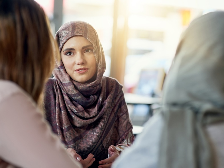 En kvinna i hijab samtalar med två andra kvinnor i ett ljust café eller mötesrum.