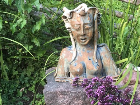 Keramikskulptur av en kvinna med långt hår står på en sten omgiven av gröna växter och lila blommor.