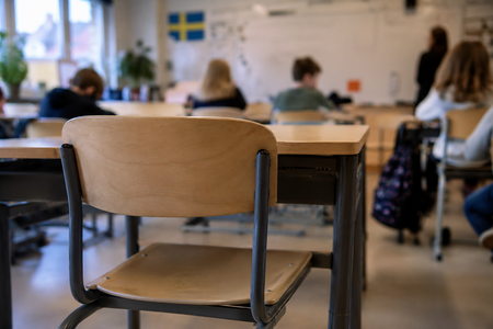Ett klassrum med tomma och upptagna skolbänkar, elever och en lärare syns suddigt i bakgrunden.