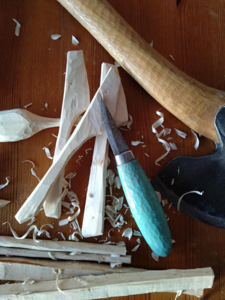 En kniv, en yxa och träbitar med spån på ett träbord, visar träslöjd i arbete.