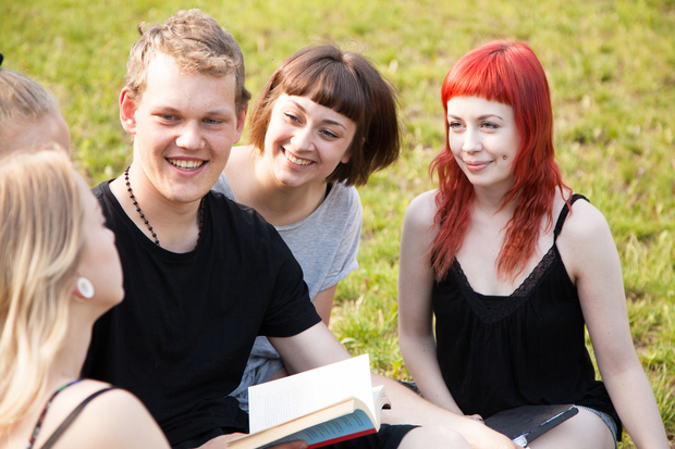 Fyra unga vuxna sitter på gräset och pratar och skrattar tillsammans utomhus.