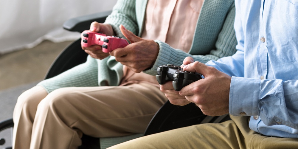 En äldre kvinna i rullstol och en yngre man spelar tv-spel tillsammans i ett ljust rum.